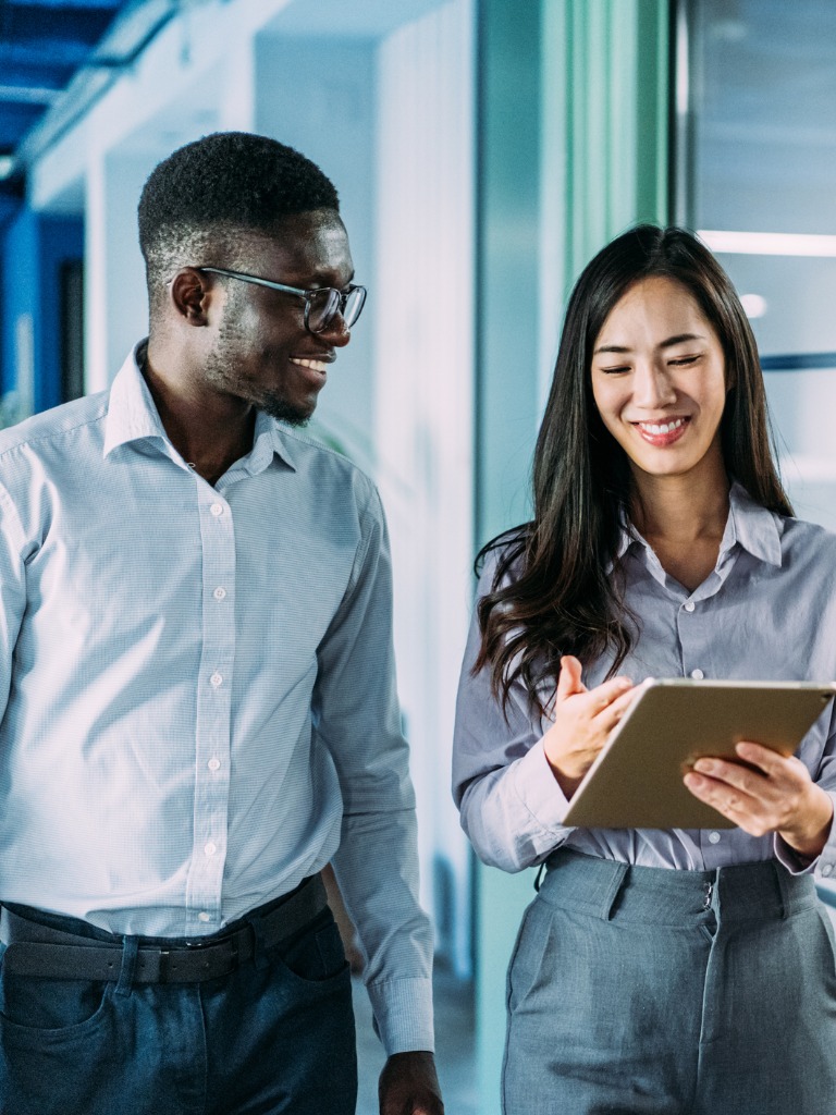 Two colleagues, a Black man and an Asian woman, smile while discussing a tablet in a modern office hallway.