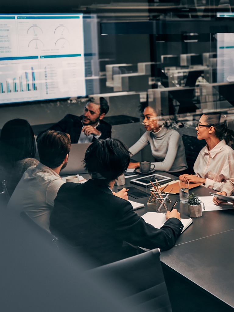 Business meeting with diverse team discussing charts on a large screen and taking notes.